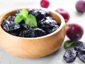 Un bol en bois rempli de pruneaux, parfaits pour lutter contre la constipation. Quelques prunes fraîches et des feuilles de menthe complètent la scène.