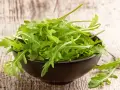 Un bol noir déborde de roquette salade fraîche, ses feuilles vertes vives contrastant avec le bois rustique.