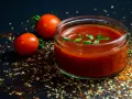 Un pot de jus de tomate frais, garni de basilic, avec deux tomates entières et des épices sur fond sombre.