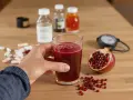 Main d'une personne tenant un verre de jus de grenade, avec une moitié de grenade, des pilules et des flacons en arrière-plan. Le jus de grenade est connu pour ses nombreux bienfaits.