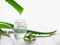 Goutte de gel d'aloe vera tombant dans un petit flacon, entouré de feuilles d'aloe vera coupées.
