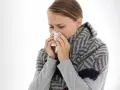 Une jeune femme enroulée dans une écharpe se mouche avec un mouchoir. Elle semble se demander combien de temps dure un rhume.