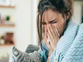 Femme enroulée dans une couverture bleue, se mouchant dans un mouchoir. Elle cherche un remède, peut-être un expectorant naturel.