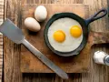 Deux œufs au plat dans une poêle, deux œufs entiers à côté. Une spatule et une salière complètent la scène. Combien d'oeuf par jour pour un bon petit déjeuner ?