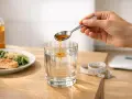 Une cuillère verse du vinaigre de cidre dans un verre d'eau. À côté, un plat de saumon et des légumes, une balance et un mètre ruban suggèrent un repas sain.