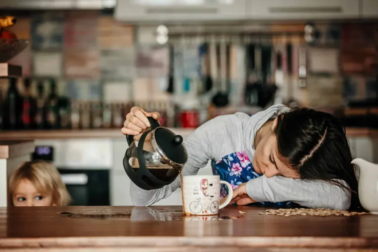 Une femme, accablée par la fatigue musculaire, verse du café dans une tasse. Un enfant la regarde, curieux.