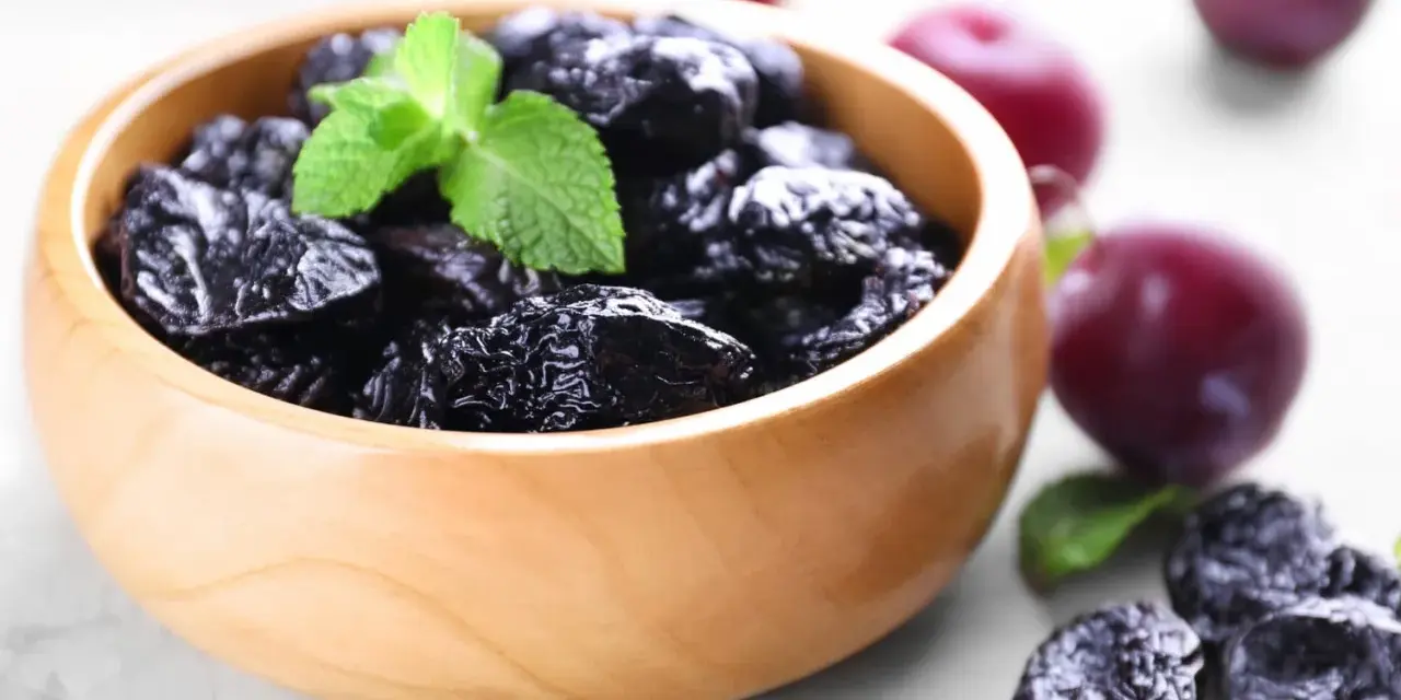 Un bol en bois rempli de pruneaux, parfaits pour lutter contre la constipation. Quelques prunes fraîches et des feuilles de menthe complètent la scène.