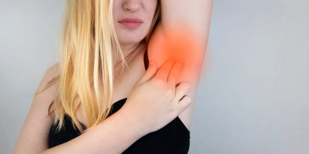 Une femme blonde exprime une douleur vive à l'aisselle, se tenant le bras avec une expression de souffrance.