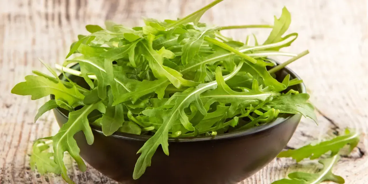 Un bol noir déborde de roquette salade fraîche, ses feuilles vertes vives contrastant avec le bois rustique.