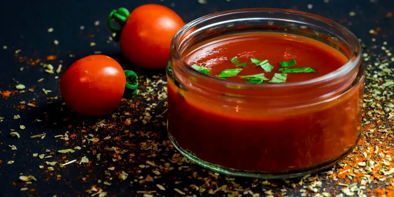 Un pot de jus de tomate frais, garni de basilic, avec deux tomates entières et des épices sur fond sombre.