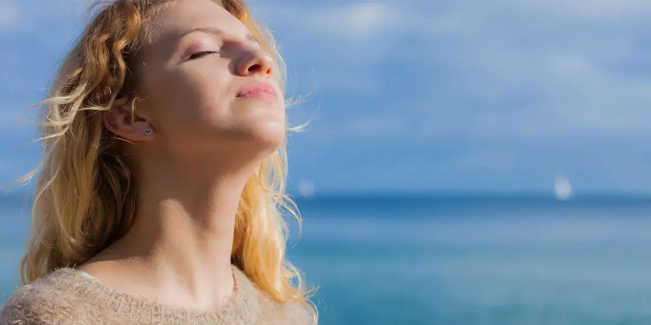 Jeune femme blonde profitant du soleil, source naturelle de vitamine D2 et D3, avec la mer en arrière-plan.