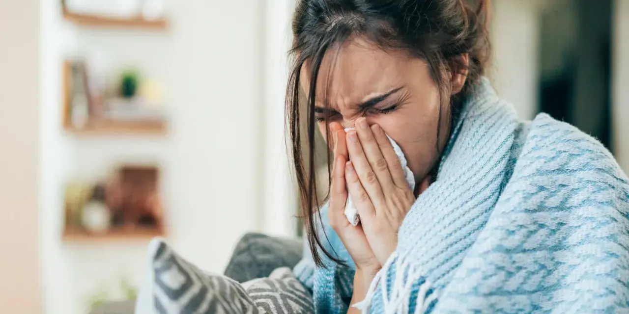 Femme enroulée dans une couverture bleue, se mouchant dans un mouchoir. Elle cherche un remède, peut-être un expectorant naturel.