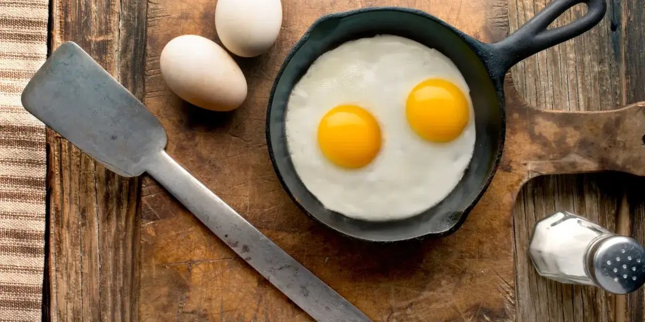 Deux œufs au plat dans une poêle, deux œufs entiers à côté. Une spatule et une salière complètent la scène. Combien d'oeuf par jour pour un bon petit déjeuner ?