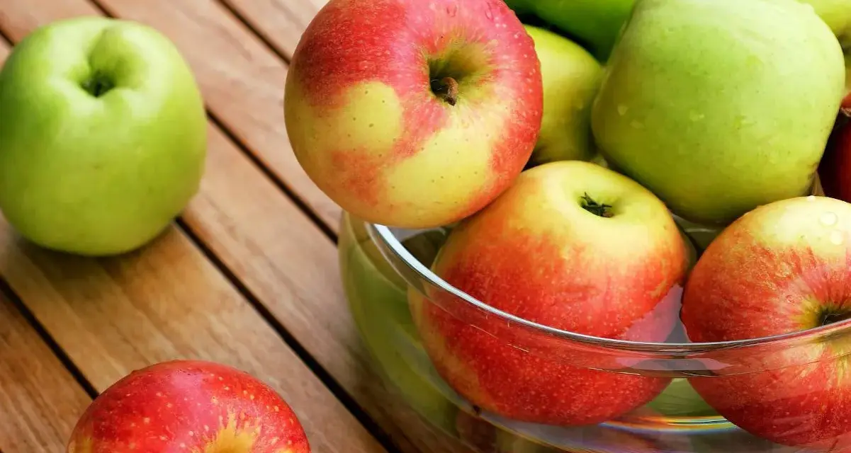 Une pomme verte et des pommes rouges et jaunes dans un bol en verre sur une table en bois.