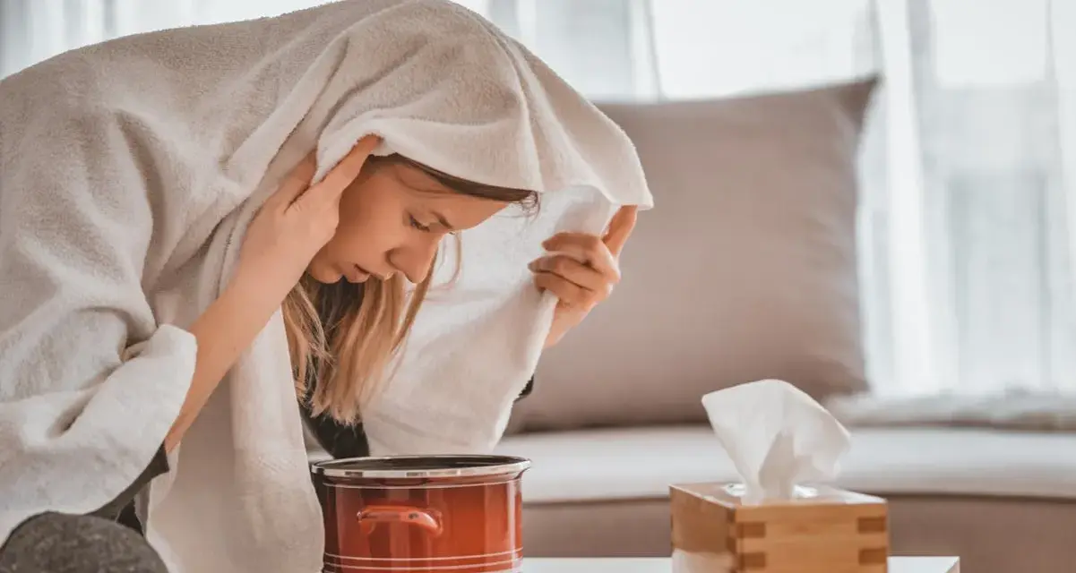 Jeune femme enroulée dans une serviette, profitant d'une inhalation vapeur pour soulager son rhume. Un pot rouge et une boîte de mouchoirs sont à proximité.