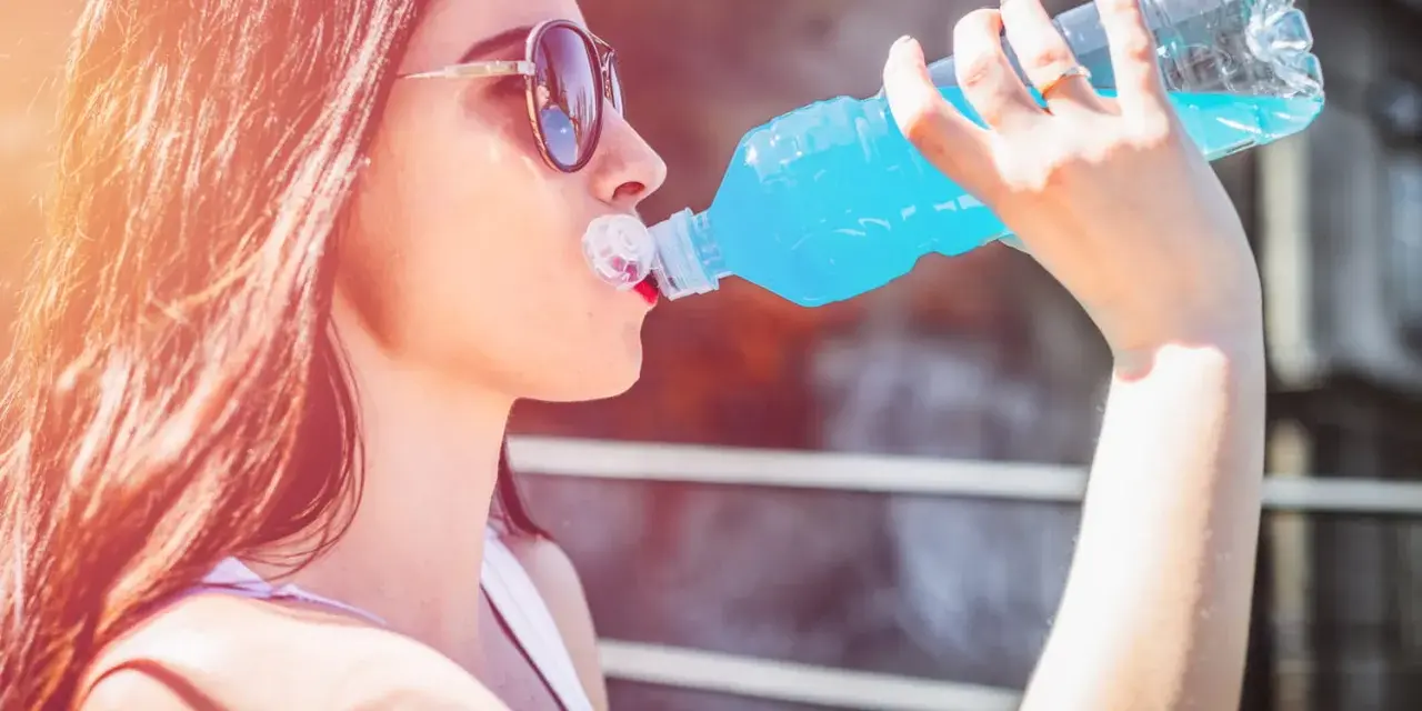 Une jeune femme aux lunettes de soleil boit une boisson bleue rafraîchissante, peut-être de l'eau de Quinton, sous le soleil.