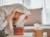 Jeune femme enroulée dans une serviette, profitant d'une inhalation vapeur pour soulager son rhume. Un pot rouge et une boîte de mouchoirs sont à proximité.