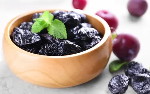 Un bol en bois rempli de pruneaux, parfaits pour lutter contre la constipation. Quelques prunes fraîches et des feuilles de menthe complètent la scène.