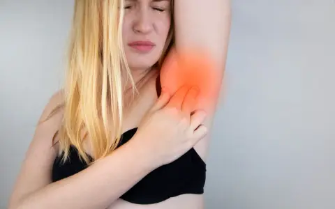 Une femme blonde exprime une douleur vive à l'aisselle, se tenant le bras avec une expression de souffrance.