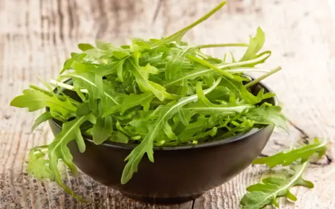Un bol noir déborde de roquette salade fraîche, ses feuilles vertes vives contrastant avec le bois rustique.