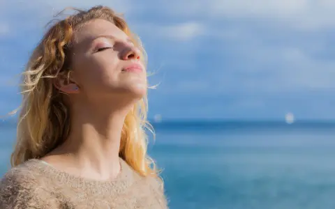 Jeune femme blonde profitant du soleil, source naturelle de vitamine D2 et D3, avec la mer en arrière-plan.