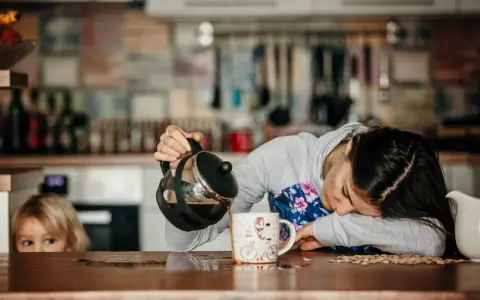 Une femme, accablée par la fatigue musculaire, verse du café dans une tasse. Un enfant la regarde, curieux.