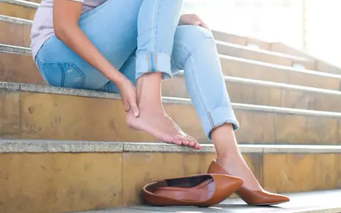 Douleur au talon ? Une femme s'assoit sur des marches, massant son pied. Peut-être un remède de grand-mère pour soulager cette gêne.