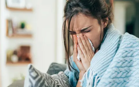 Femme enroulée dans une couverture bleue, se mouchant dans un mouchoir. Elle cherche un remède, peut-être un expectorant naturel.