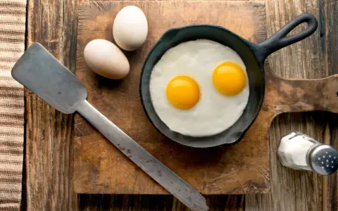 Deux œufs au plat dans une poêle, deux œufs entiers à côté. Une spatule et une salière complètent la scène. Combien d'oeuf par jour pour un bon petit déjeuner ?