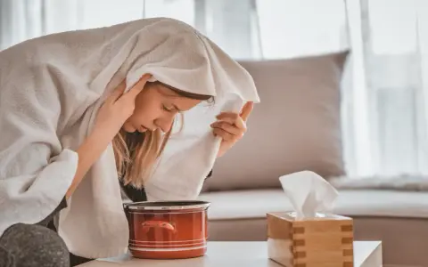 Jeune femme enroulée dans une serviette, profitant d'une inhalation vapeur pour soulager son rhume. Un pot rouge et une boîte de mouchoirs sont à proximité.
