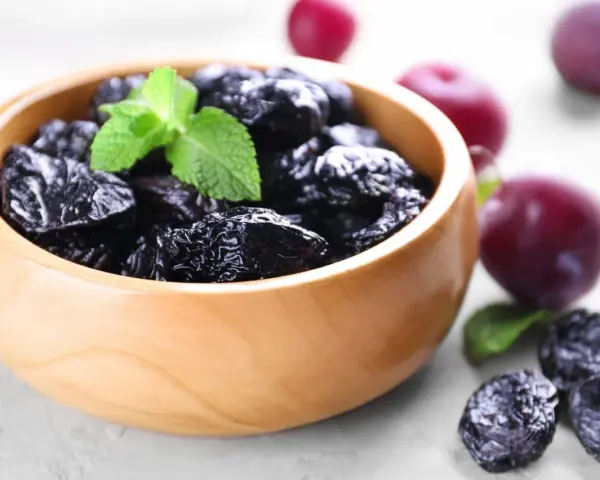 Un bol en bois rempli de pruneaux, parfaits pour lutter contre la constipation. Quelques prunes fraîches et des feuilles de menthe complètent la scène.
