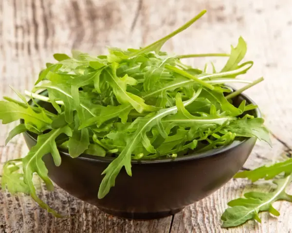 Un bol noir déborde de roquette salade fraîche, ses feuilles vertes vives contrastant avec le bois rustique.