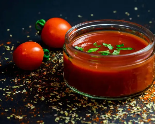 Un pot de jus de tomate frais, garni de basilic, avec deux tomates entières et des épices sur fond sombre.