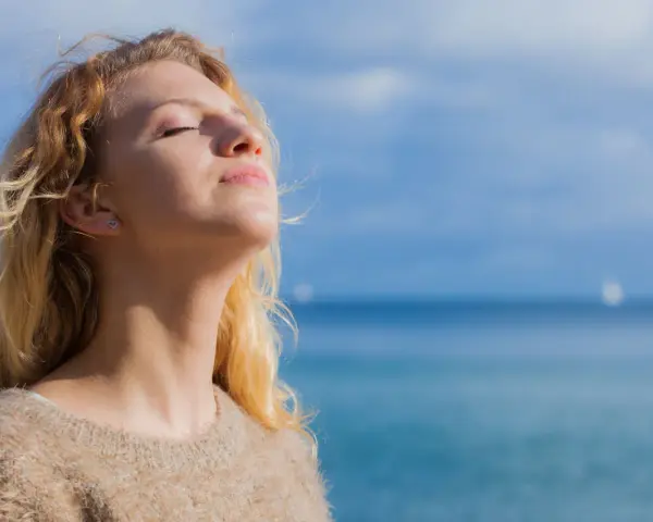 Jeune femme blonde profitant du soleil, source naturelle de vitamine D2 et D3, avec la mer en arrière-plan.