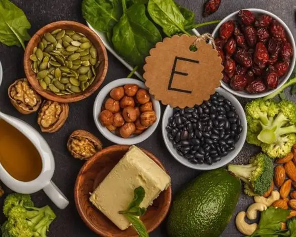 Assortiment d'aliments riches en vitamine E : amandes, noisettes, épinards, avocat, haricots noirs, graines de courge, et huile.