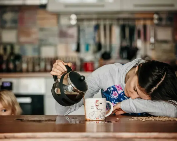 Une femme, accablée par la fatigue musculaire, verse du café dans une tasse. Un enfant la regarde, curieux.