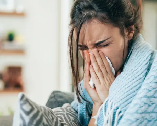 Femme enroulée dans une couverture bleue, se mouchant dans un mouchoir. Elle cherche un remède, peut-être un expectorant naturel.