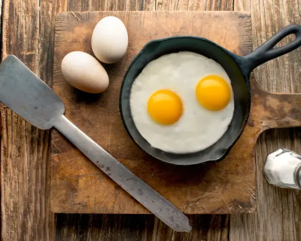 Deux œufs au plat dans une poêle, deux œufs entiers à côté. Une spatule et une salière complètent la scène. Combien d'oeuf par jour pour un bon petit déjeuner ?