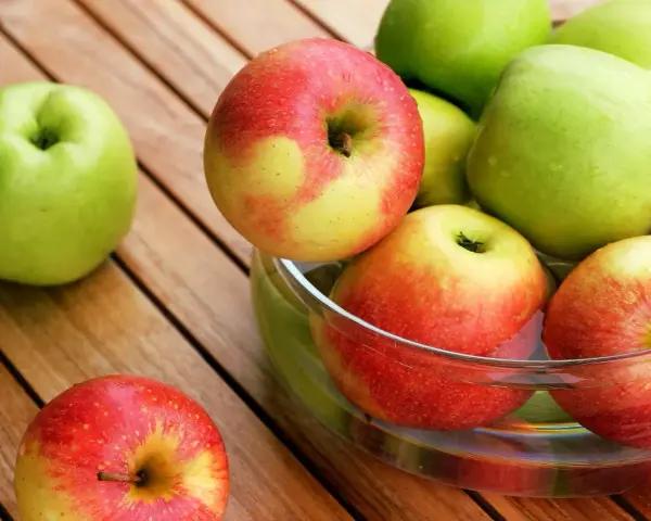 Une pomme verte et des pommes rouges et jaunes dans un bol en verre sur une table en bois.