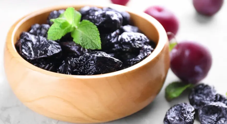Un bol en bois rempli de pruneaux, parfaits pour lutter contre la constipation. Quelques prunes fraîches et des feuilles de menthe complètent la scène.