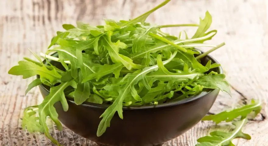 Un bol noir déborde de roquette salade fraîche, ses feuilles vertes vives contrastant avec le bois rustique.