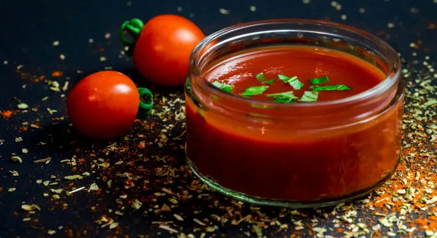 Un pot de jus de tomate frais, garni de basilic, avec deux tomates entières et des épices sur fond sombre.