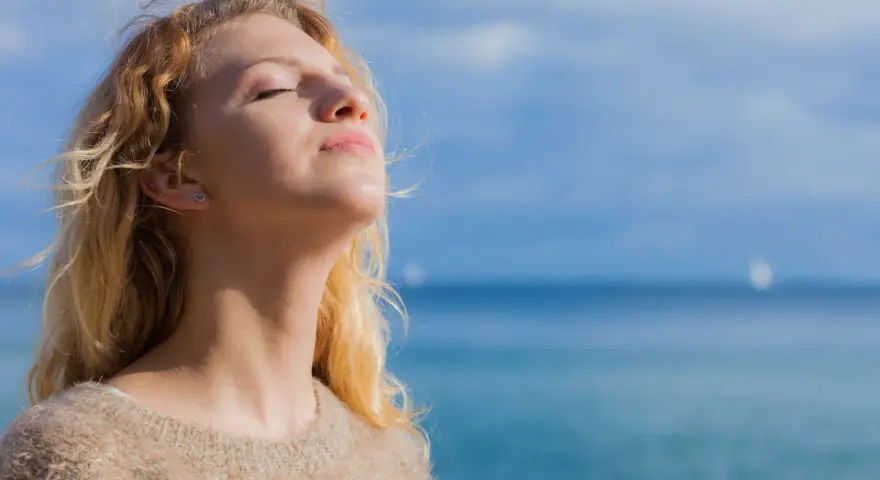 Jeune femme blonde profitant du soleil, source naturelle de vitamine D2 et D3, avec la mer en arrière-plan.