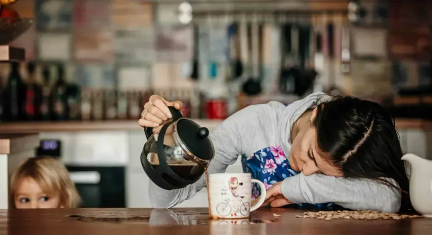 Une femme, accablée par la fatigue musculaire, verse du café dans une tasse. Un enfant la regarde, curieux.