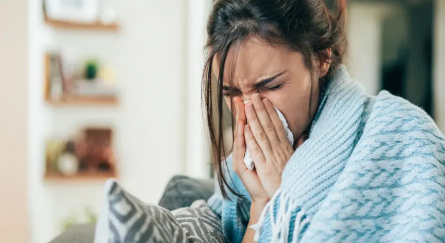 Femme enroulée dans une couverture bleue, se mouchant dans un mouchoir. Elle cherche un remède, peut-être un expectorant naturel.