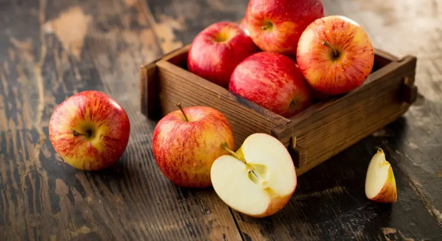 Des pommes rouges et jaunes dans une caisse en bois sur une table rustique. Une pomme est coupée en deux, révélant sa chair blanche. Ces fruits sont un concentré de pomme bienfaits.
