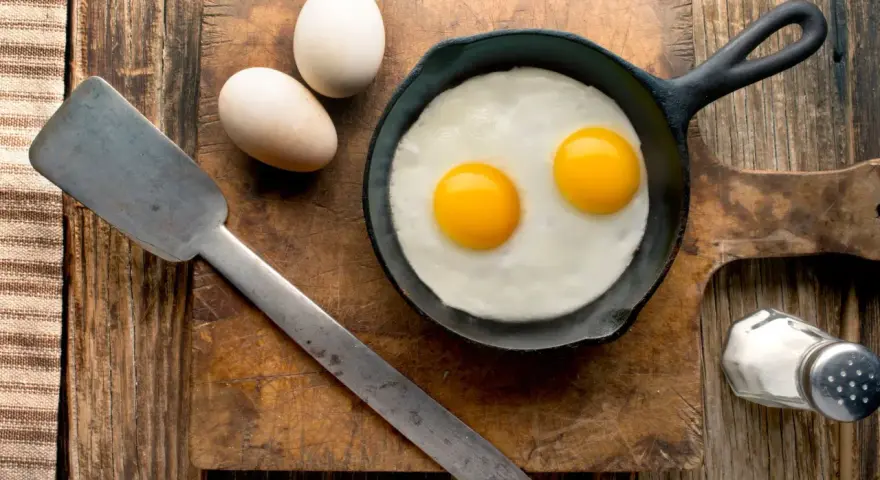 Deux œufs au plat dans une poêle, deux œufs entiers à côté. Une spatule et une salière complètent la scène. Combien d'oeuf par jour pour un bon petit déjeuner ?