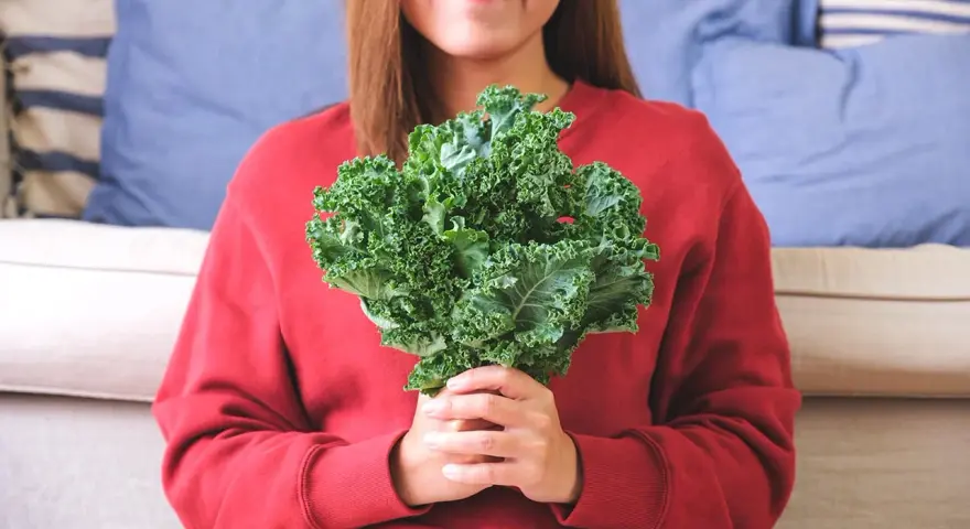 Une femme souriante tient un bouquet de chou kale, un légume aux multiples chou kale bienfaits pour la santé.