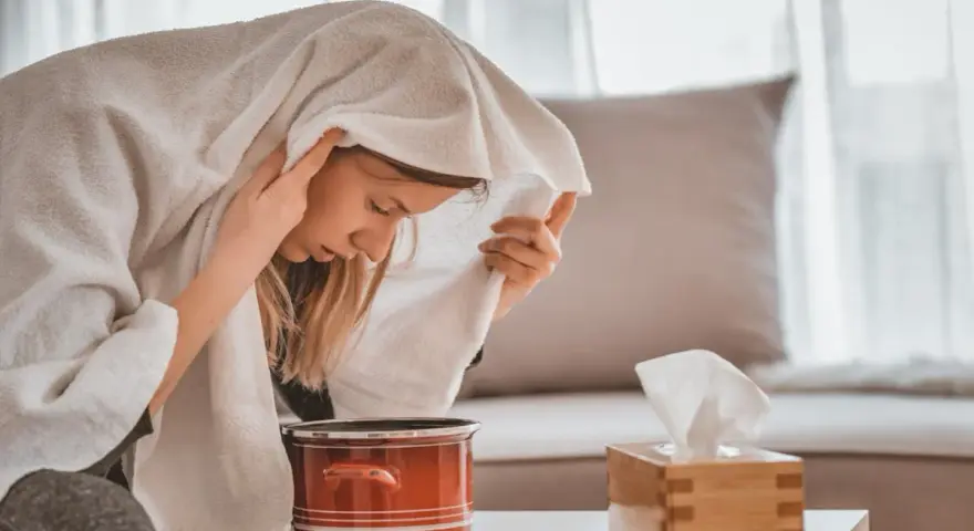 Jeune femme enroulée dans une serviette, profitant d'une inhalation vapeur pour soulager son rhume. Un pot rouge et une boîte de mouchoirs sont à proximité.