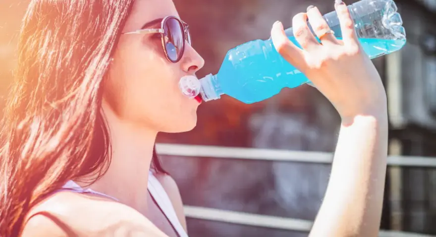 Une jeune femme aux lunettes de soleil boit une boisson bleue rafraîchissante, peut-être de l'eau de Quinton, sous le soleil.